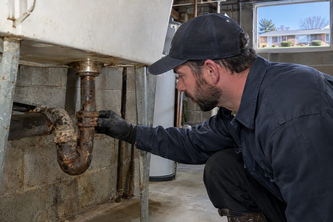 Close view of a plumber inspecting a corroded cast iron drain beneath a basement utility sink in an established Dayton-area home in West Carrollton, OH